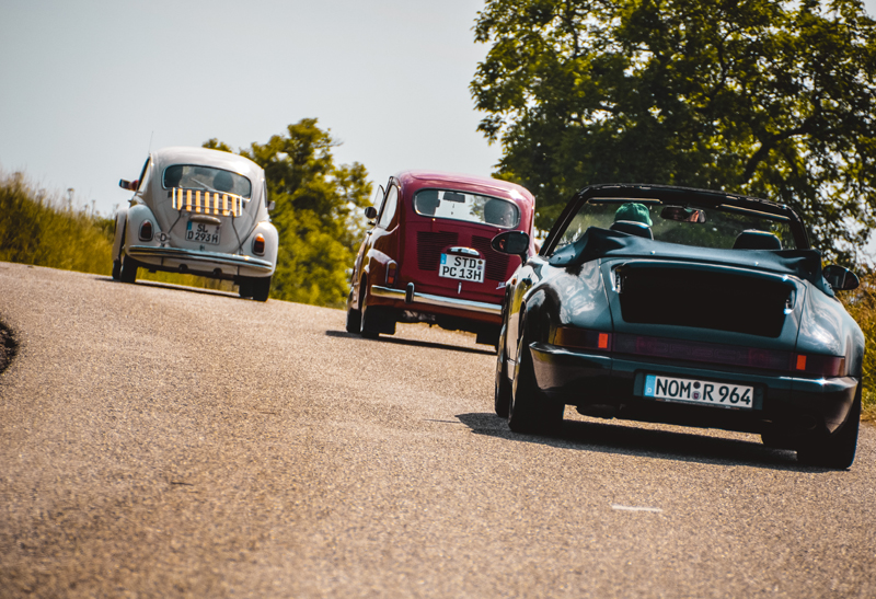 Ein alter Porsche sowie zwei alte VW Käfer auf der Straße während einer Tour über die Landstraße